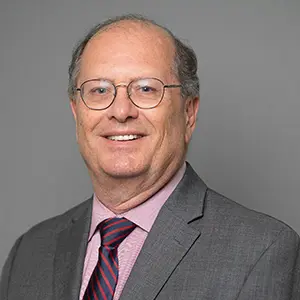 Kevin Shoemaker in a gray suit with a pink shirt and striped tie, smiling in front of a neutral gray background.