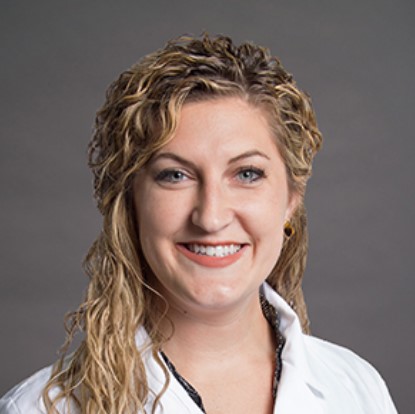 Good Dr. Good wearing a white coat, smiling with wavy blonde hair, in front of a dark gray background.