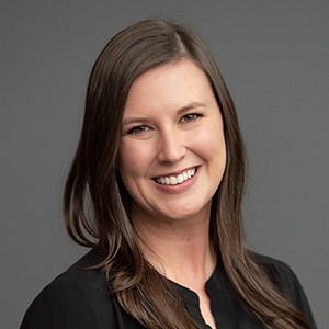 Dr. Maxwell smiling in a black blouse with long brown hair against a gray background.