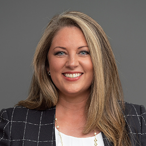 Holly Haynes wearing a black checked blazer and white blouse smiles during a professional headshot against a plain gray background.